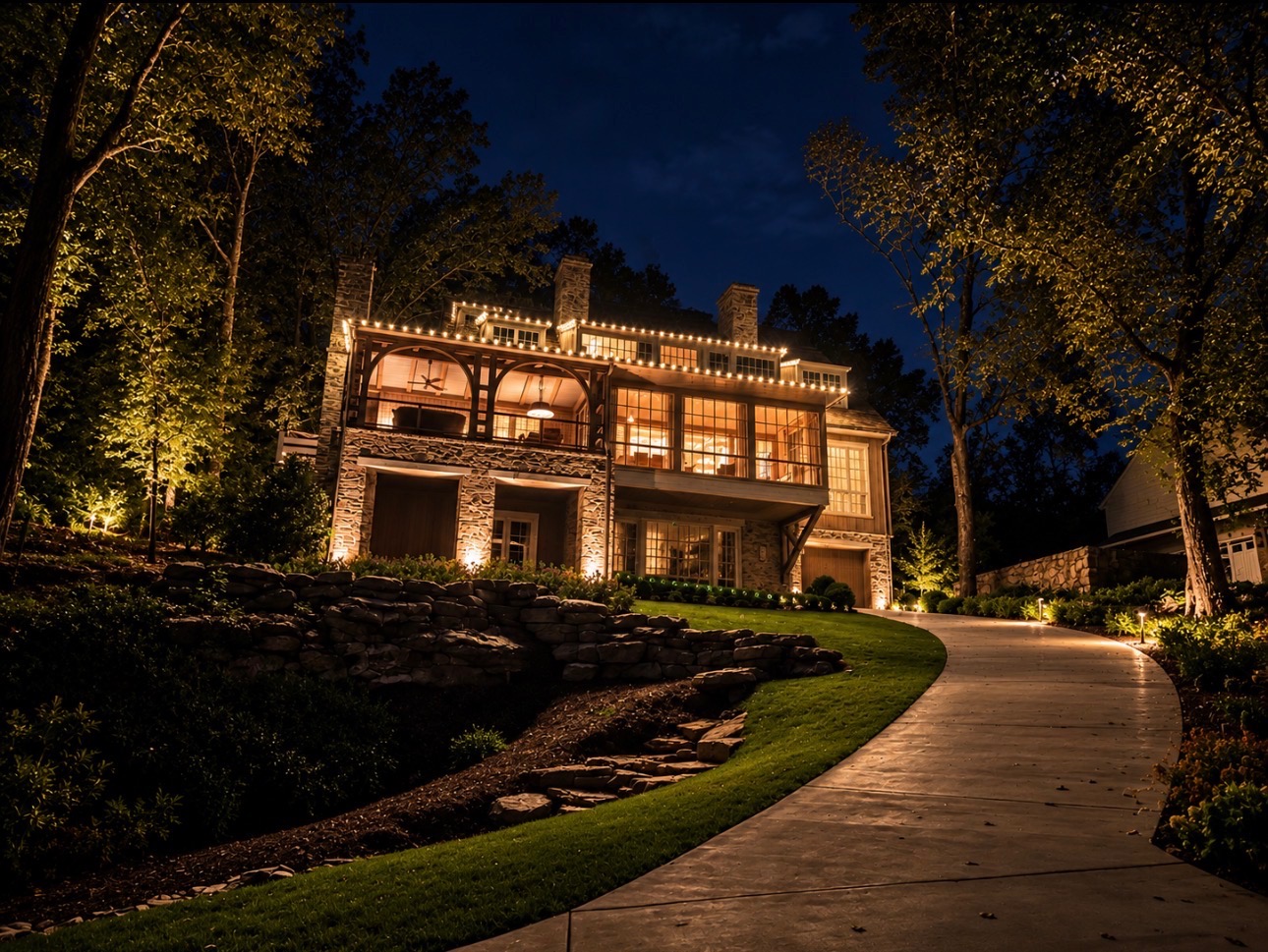 Large hillside home at night with warm white Christmas lights along the upper roofline, mature trees uplit in gold, and a curved driveway edged with warm path lights